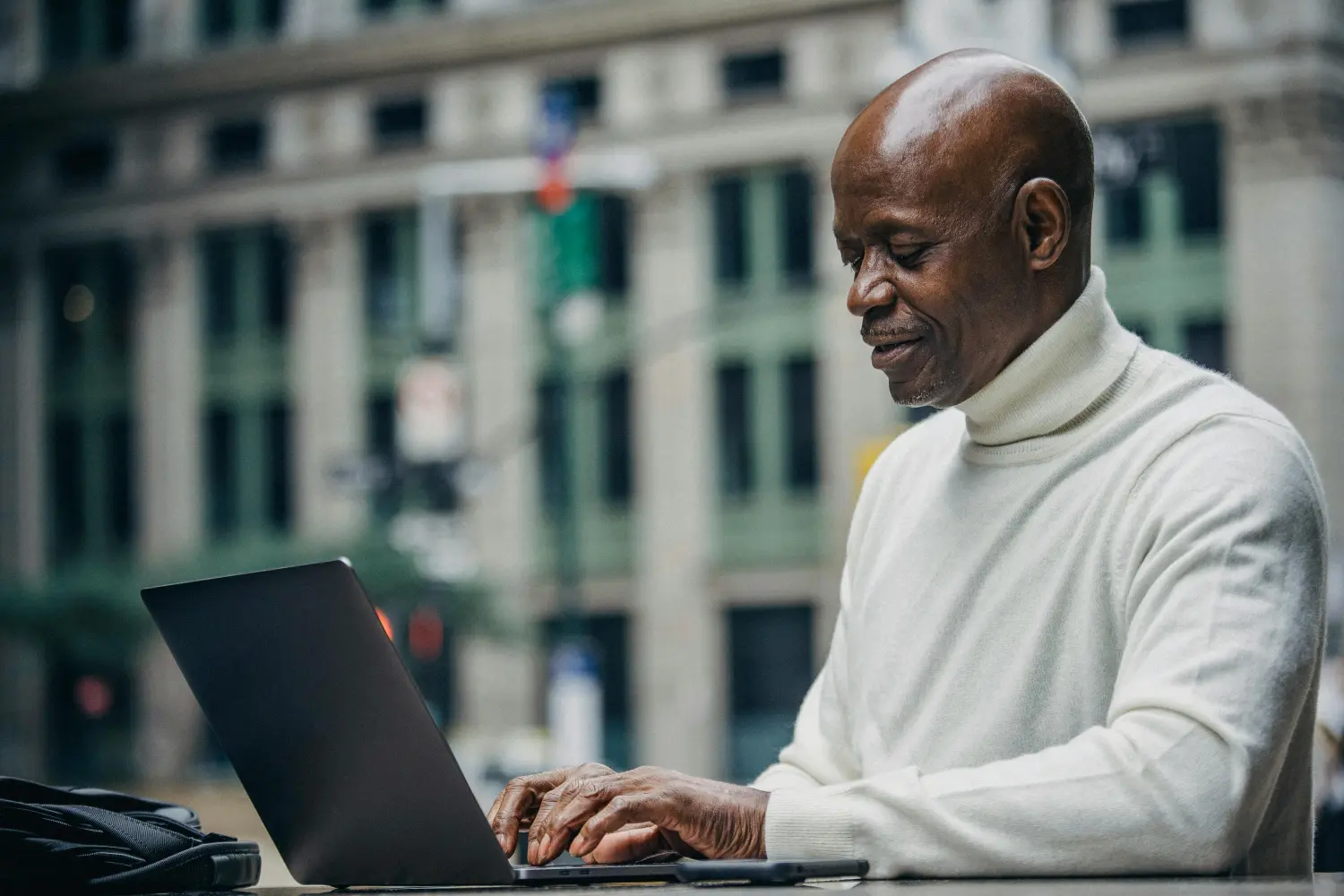 Story Notes Guy Best An older man sits typing on his laptop at an outdoor cafe in a city.